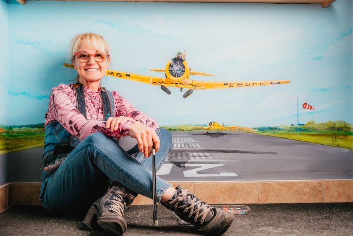 Photo of local artist Stella Jurgen in front of the Skyway Café featuring a hand-painted iconic yellow Harvard taking off from the Tillsonburg Airport runway. Photo courtesy of Oxford County Tourism and Dudek Photography.